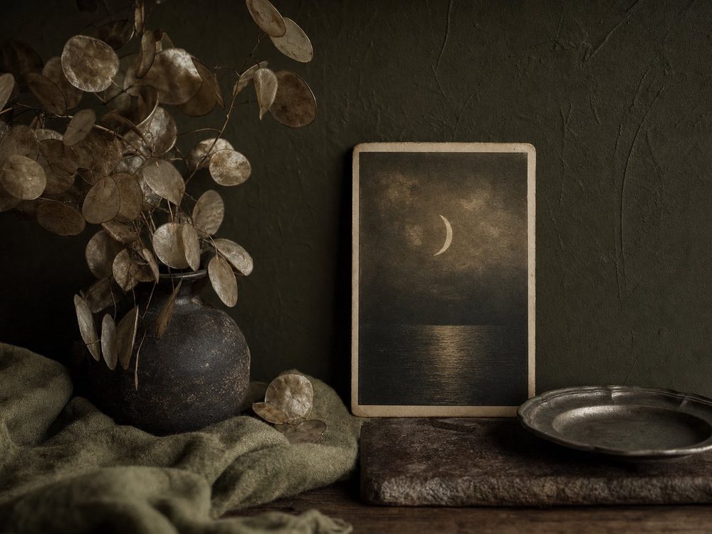 A framed antique card print of a moon above a narrow path, beside dried stems in a dark vessel on a stone shelf.